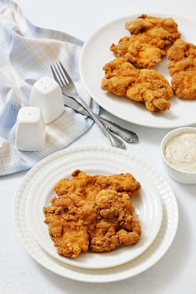 Plate of golden fried chicken on a white table, flanked by a checkered napkin, utensils, salt shakers, and a small dish of dipping sauce. Warm, inviting tone.
