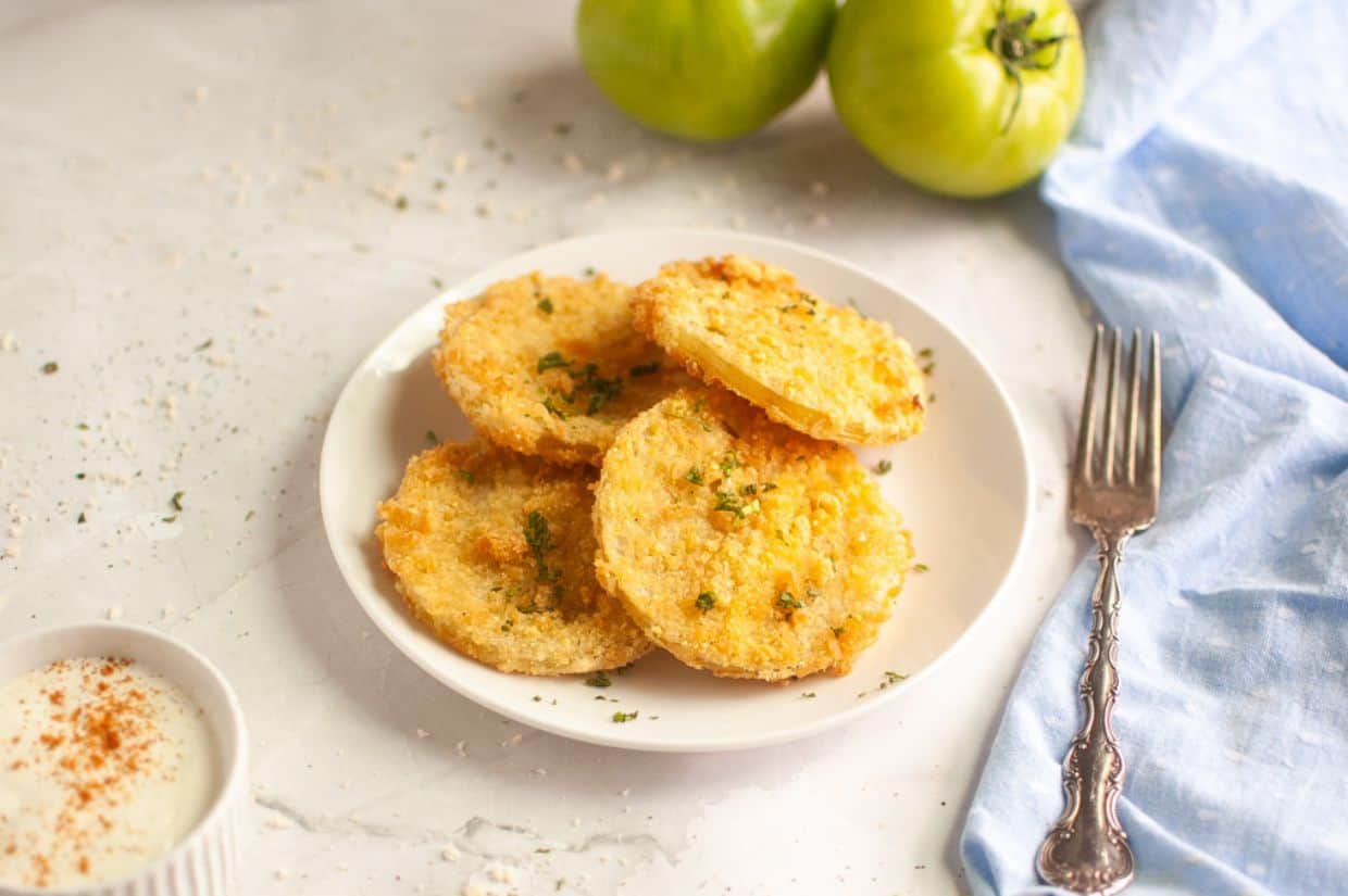 Plate of crispy, golden fried green tomatoes garnished with herbs. Beside them are two fresh green tomatoes and a blue napkin with a fork.