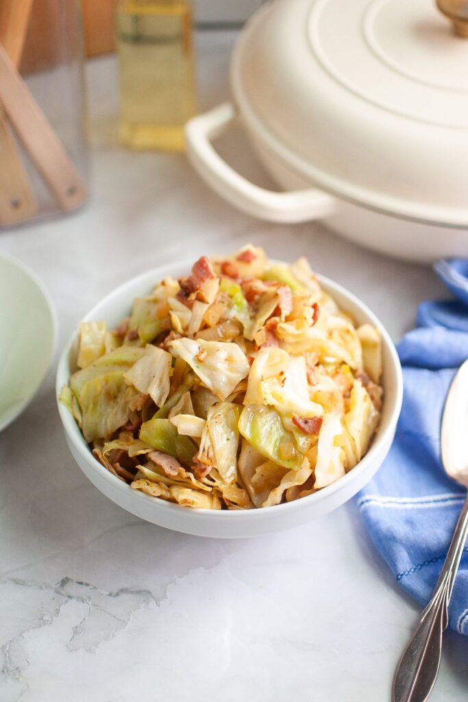 Bowl of fried cabbage with bacon on a marble countertop. A white pot and blue napkin with a spoon are nearby, creating a cozy kitchen scene.