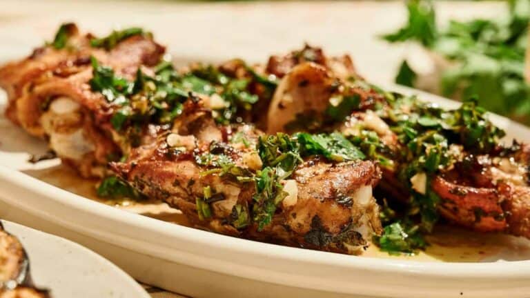 Grilled meat ribs garnished with chopped herbs and garlic, served on a white platter with fresh parsley in the background.