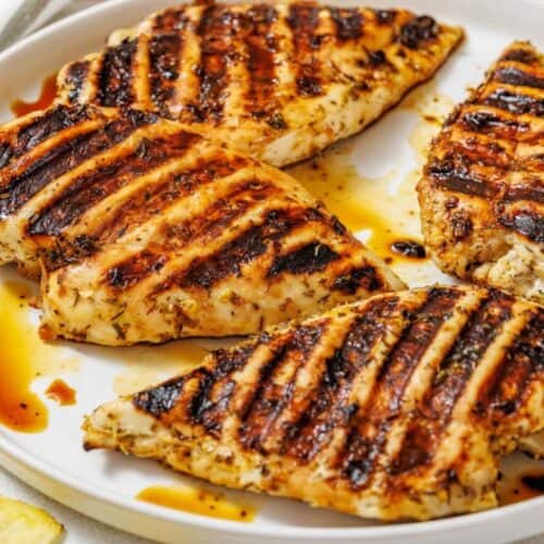 Grilled chicken breasts with charred grill marks, drizzled with sauce on a white plate, exuding a savory aroma.