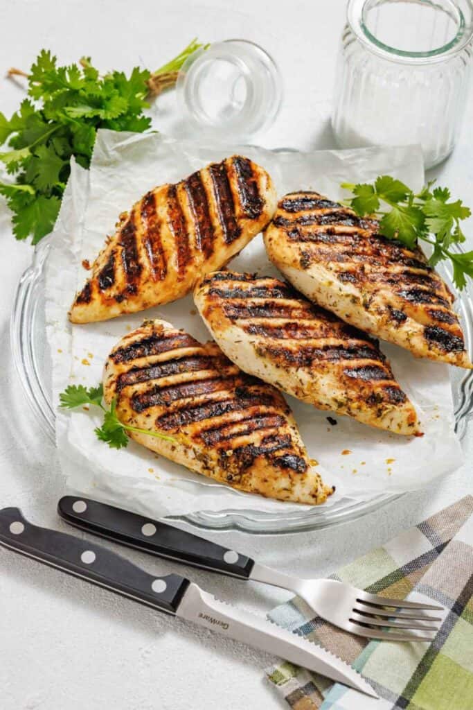 Grilled chicken breasts with char marks on parchment-lined glass plate. Garnished with cilantro, surrounded by parsley, knife, fork, and salt.