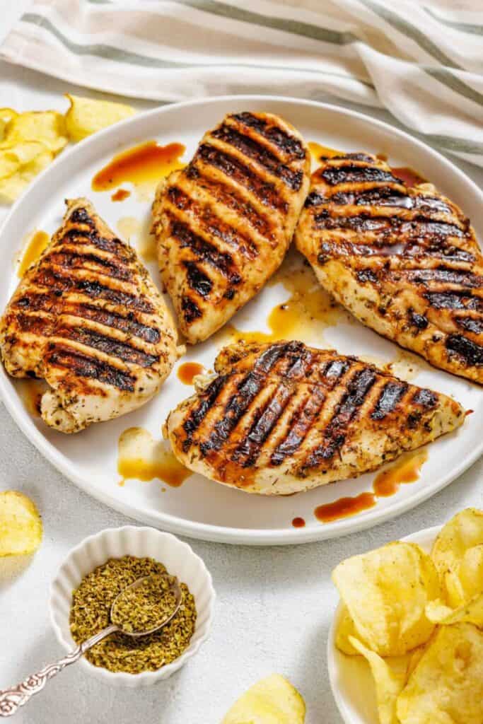 Grilled chicken breasts with char marks on a white plate, drizzled with sauce. Nearby are potato chips and a small bowl of herbs with a spoon.