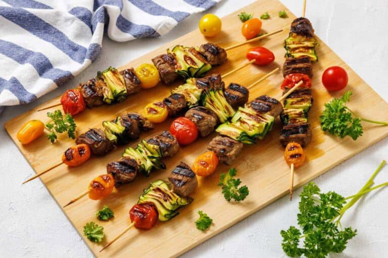 Grilled beef and vegetable skewers with cherry tomatoes, zucchini, and parsley, arranged on a wooden board next to a striped cloth.