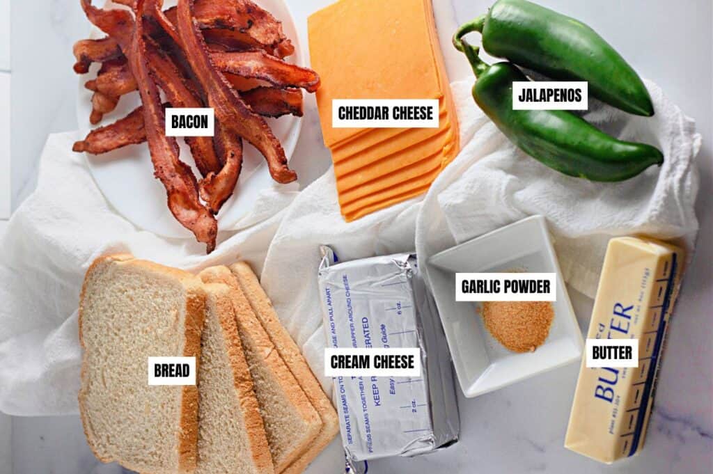 Slices of bacon, cheddar cheese, jalapeños, and bread are arranged with cream cheese, garlic powder, and butter on a white surface.