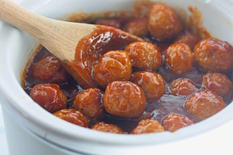 Honey-Garlic Meatballs in a thick red sauce are being stirred with a wooden spoon in a white ceramic pot.