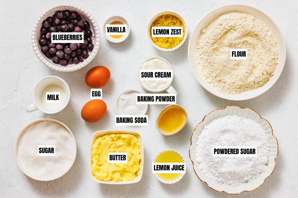 Assorted baking ingredients labeled on a white surface. Blueberries, flour, sugar, butter, eggs, milk, lemon zest, and more, evoke a fresh, homemade vibe.
