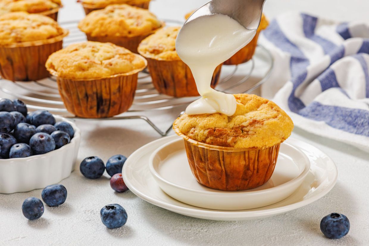 Golden muffins on a wire rack, with one on a plate being drizzled with white icing. Fresh blueberries scattered nearby, adding a fresh touch.
