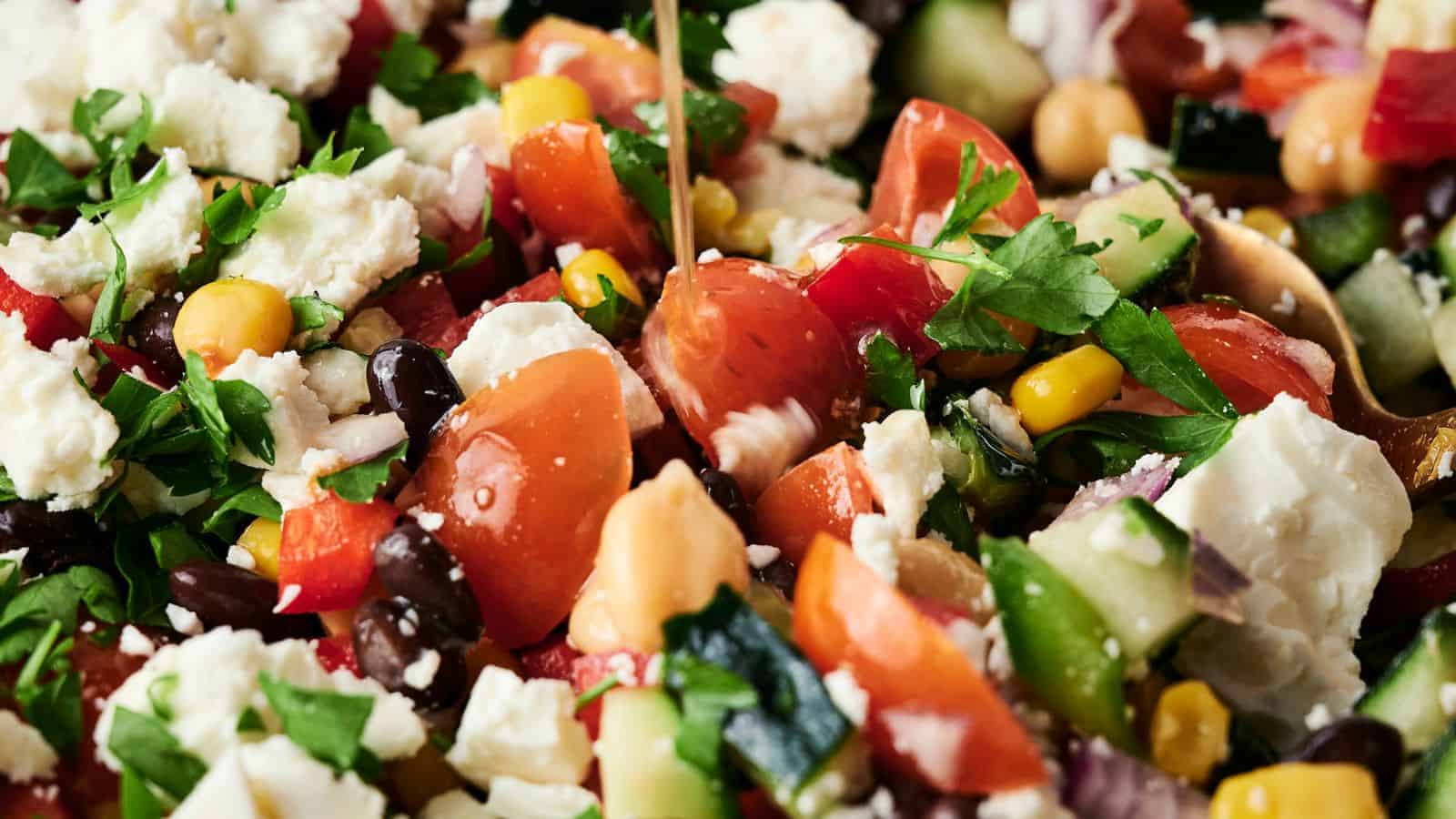Close-up of a mixed salad with tomatoes, feta cheese, black beans, chickpeas, corn, cucumber, red onion, and parsley, being drizzled with dressing.