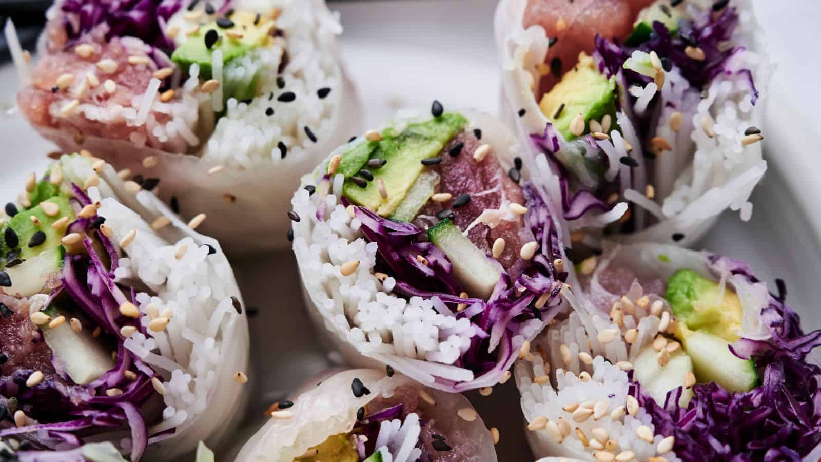 Close-up of sliced spring rolls filled with rice, avocado, purple cabbage, and sesame seeds, arranged on a white plate.