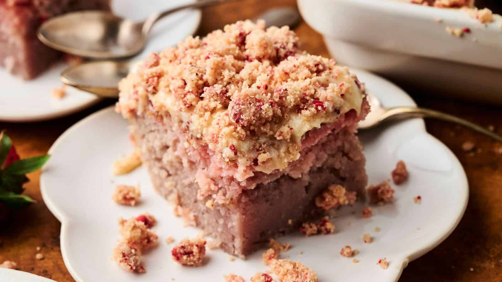 A slice of crumb-topped strawberry coffee cake sits on a white plate with a spoon beside it; additional cake and plates are in the background.