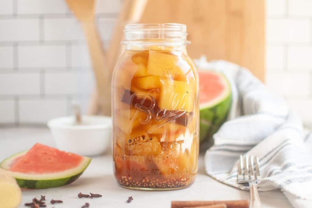 Jar of pickled watermelon rind and spices on a kitchen counter; nearby, a watermelon wedge and a striped towel enhance the fresh, rustic setting.