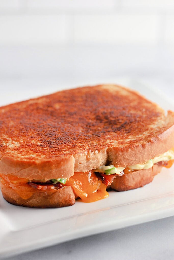 Close-up of a crispy grilled cheese sandwich on a white plate. Melted cheese oozes from the golden-brown toasted bread, creating a warm, comforting vibe.