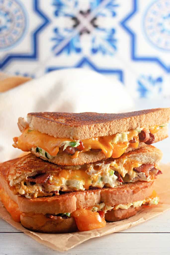 Toasted sandwich overflowing with melted cheddar, bacon, and cream cheese on brown paper.