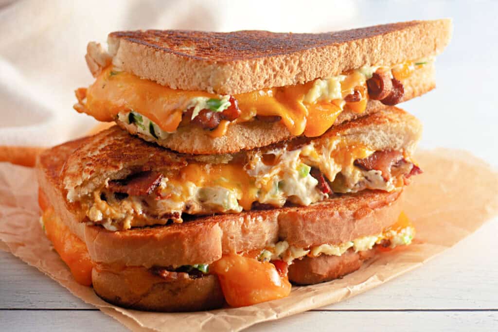 Close-up of two stacked grilled cheese sandwiches on parchment paper. Melted cheddar, bacon, and scallions ooze from crispy toasted bread, creating a warm, savory appeal.