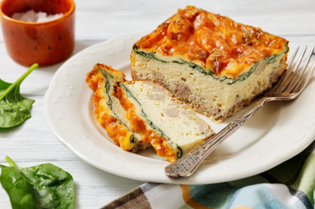 A savory breakfast casserole slice with cheese, spinach, and sausage on a white plate, accompanied by a fork, fresh spinach leaves, and a salt bowl.