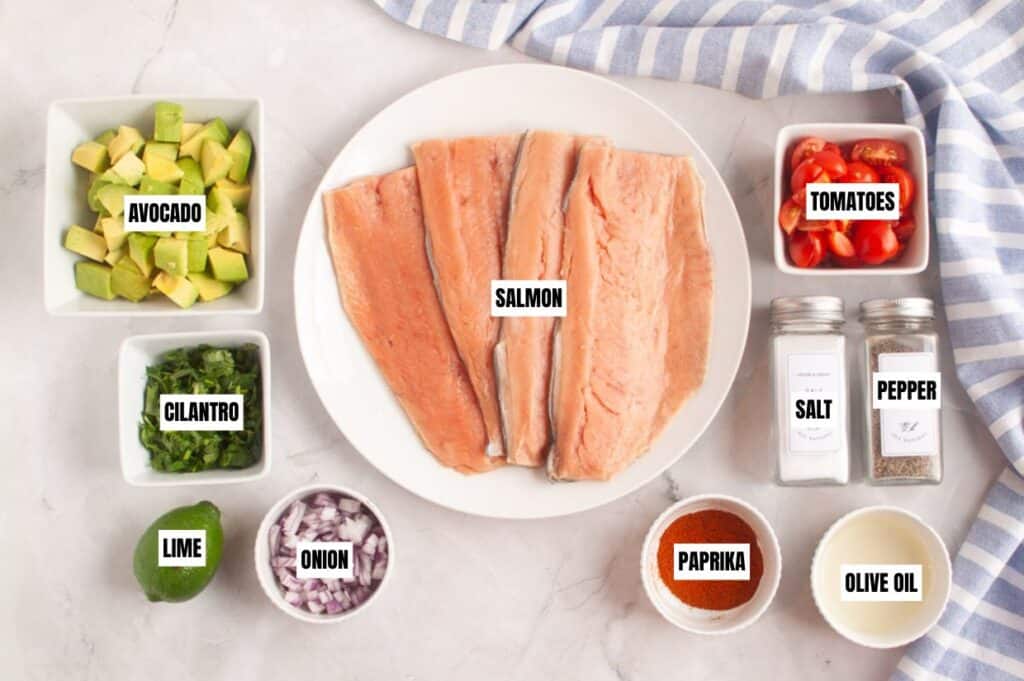 Salmon fillets on a plate surrounded by avocado, cilantro, cherry tomatoes, lime, onion, paprika, salt, pepper, and olive oil on a marble surface.