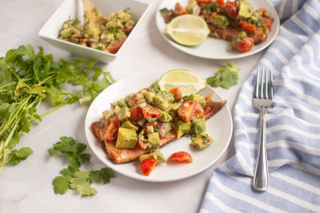 Grilled salmon topped with avocado and tomato salsa on a white plate, garnished with lime slices.