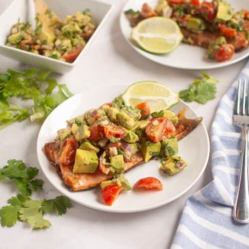 Grilled salmon topped with avocado and tomato salsa on a white plate, garnished with lime slices.