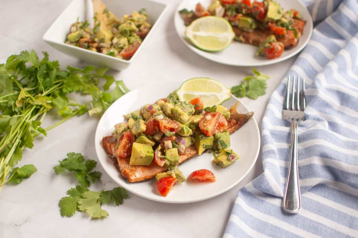 Grilled salmon topped with avocado and tomato salsa on a white plate, garnished with lime slices.