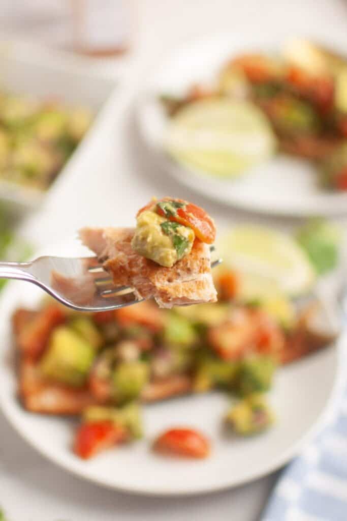 Close-up of a fork holding a piece of salmon topped with avocado salad, blurred plate in background. Fresh, vibrant, and appetizing presentation.