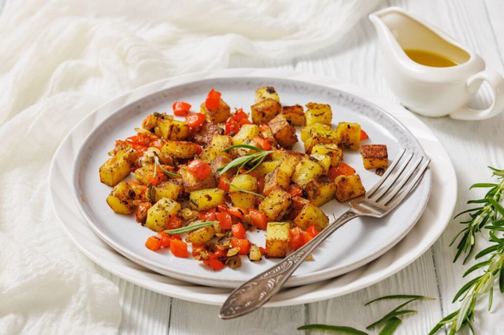 Plate of roasted potatoes with red bell peppers and herbs on a white dish, accompanied by a vintage fork. Olive oil in a gravy boat is in the background.