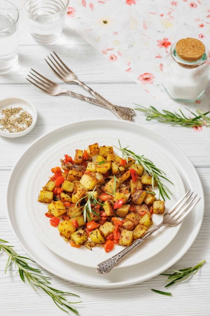 A plate of seasoned diced potatoes and red bell peppers with sprigs of rosemary on a white table. Forks, salt, and pepper add a rustic touch.