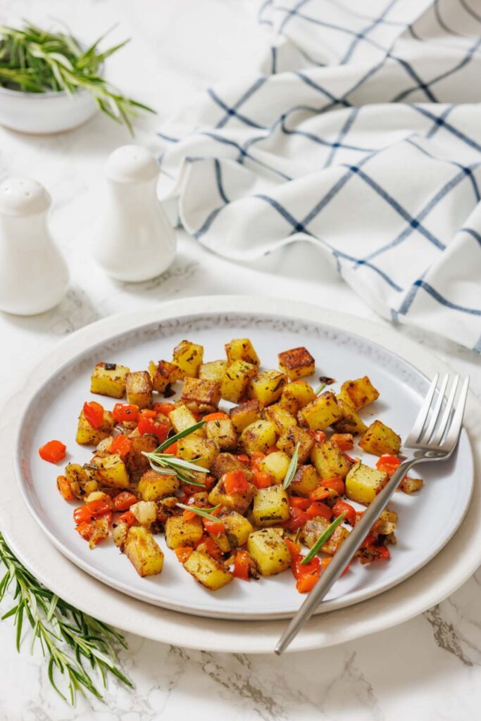 A plate of golden-brown roasted potatoes and diced red peppers garnished with rosemary, placed on a white tablecloth with a fork beside it.