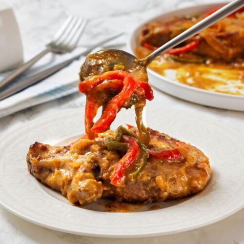 Plate of pork chops with gravy, topped with red and green bell peppers. A fork holds a bite above the dish. Background includes cutlery sand a striped napkin.