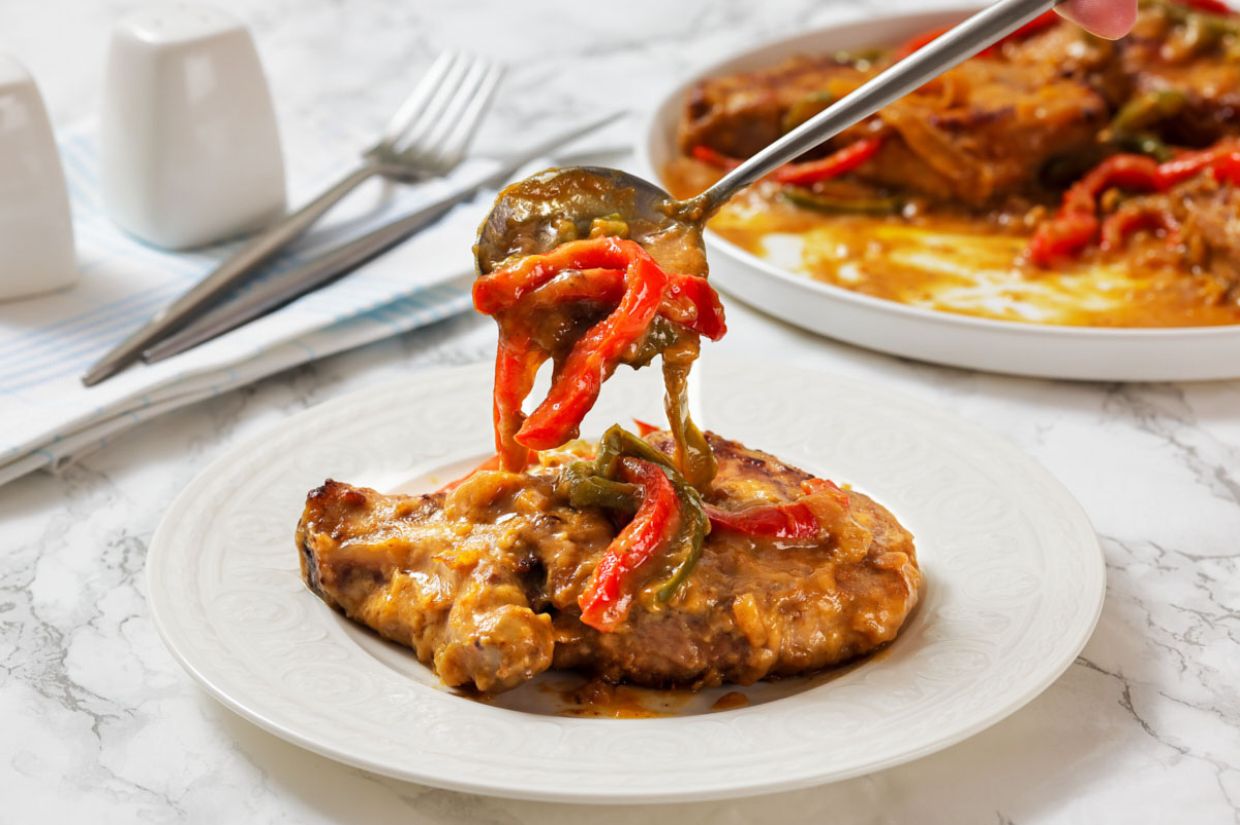 Plate of smothered pork chops with gravy, topped with red and green bell peppers. A fork holds a bite above the dish. Background includes cutlery sand a striped napkin.