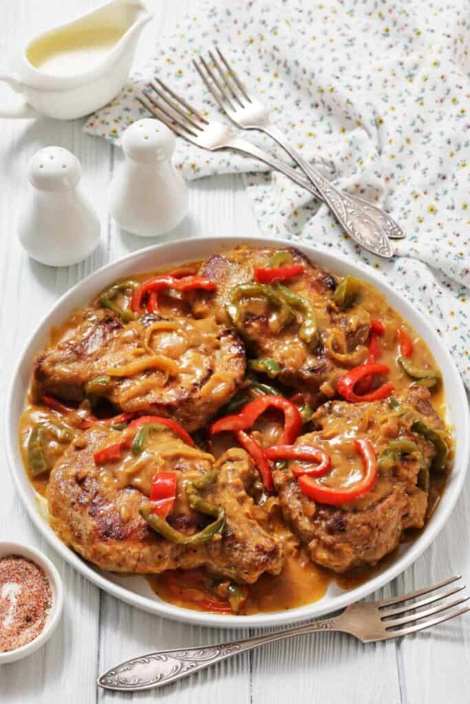 A plate of smothered pork chops with red and green bell peppers in a savory sauce. Nearby are salt shakers, a cream jug, and silver forks on a floral napkin.