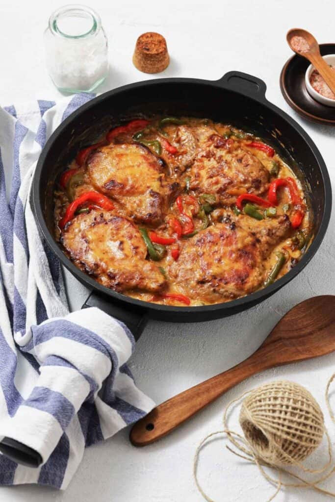 A skillet with four browned pork chops in creamy sauce, surrounded by red and green bell peppers. A striped towel, wooden spoon, salt, and a cork are nearby.
