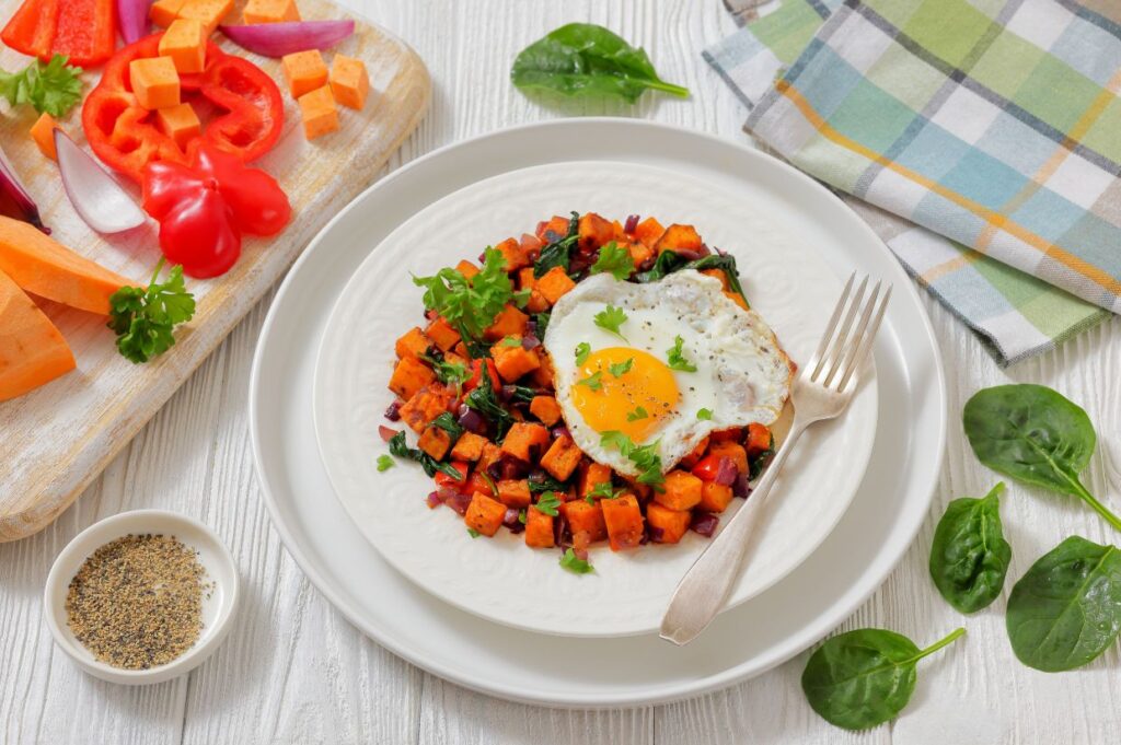 Plate of vibrant spinach sweet potato hash with a sunny-side-up egg and greens, surrounded by fresh vegetables and spices on a wooden table. Cozy and inviting.