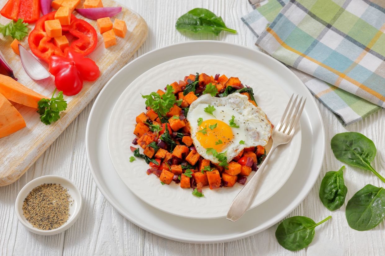 Plate of vibrant spinach sweet potato hash with a sunny-side-up egg and greens, surrounded by fresh vegetables and spices on a wooden table. Cozy and inviting.