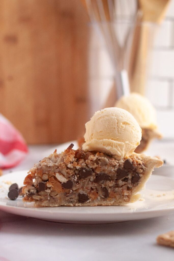 A slice of Texas Trash Pie filled with chocolate chips, pretzels, and pecans, topped with a scoop of vanilla ice cream on a white plate.