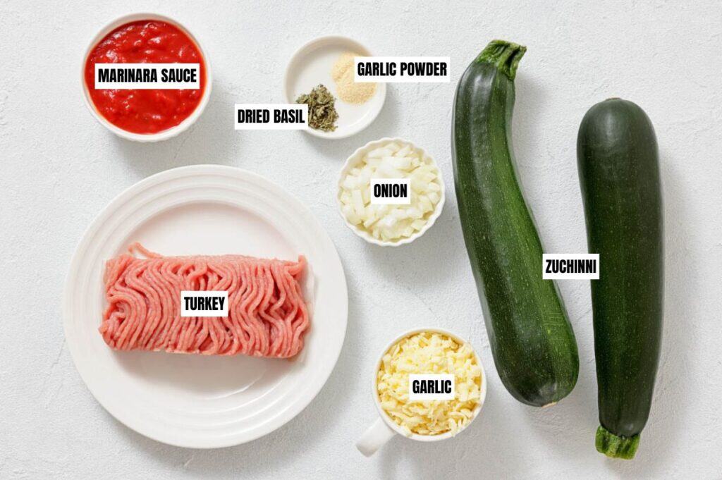 Ingredients on a white surface: ground turkey, zucchini, diced onion, minced garlic, marinara sauce, dried basil, and garlic powder. Perfect for cooking.