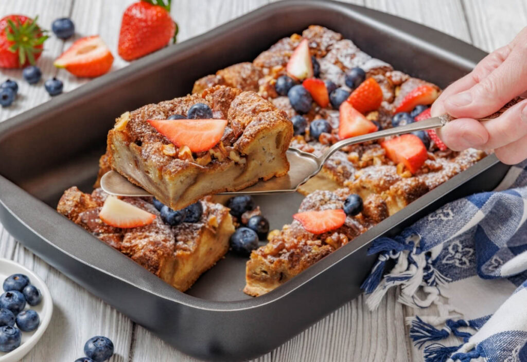 A baking pan filled with golden-brown baked cinnamon French toast, topped with fresh sliced strawberries, blueberries, and a dusting of powdered sugar.