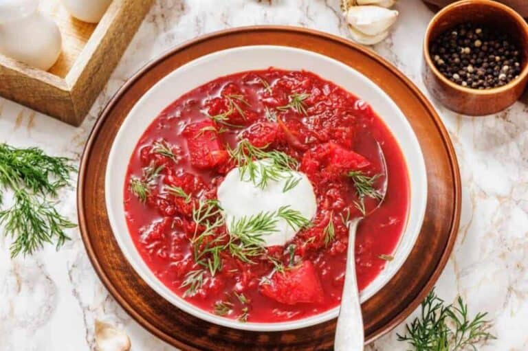 A bowl of borscht topped with a dollop of sour cream and fresh dill, served on a plate with a spoon on a marble surface.