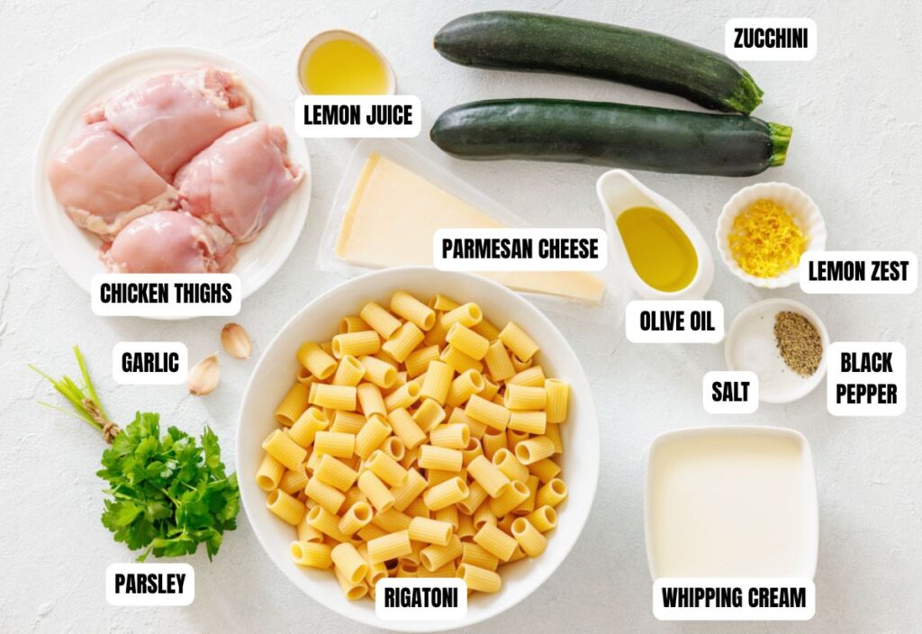 Ingredients for pasta dish are arranged on a white surface, including chicken thighs, rigatoni, zucchini, lemon juice, parmesan, and various seasonings.