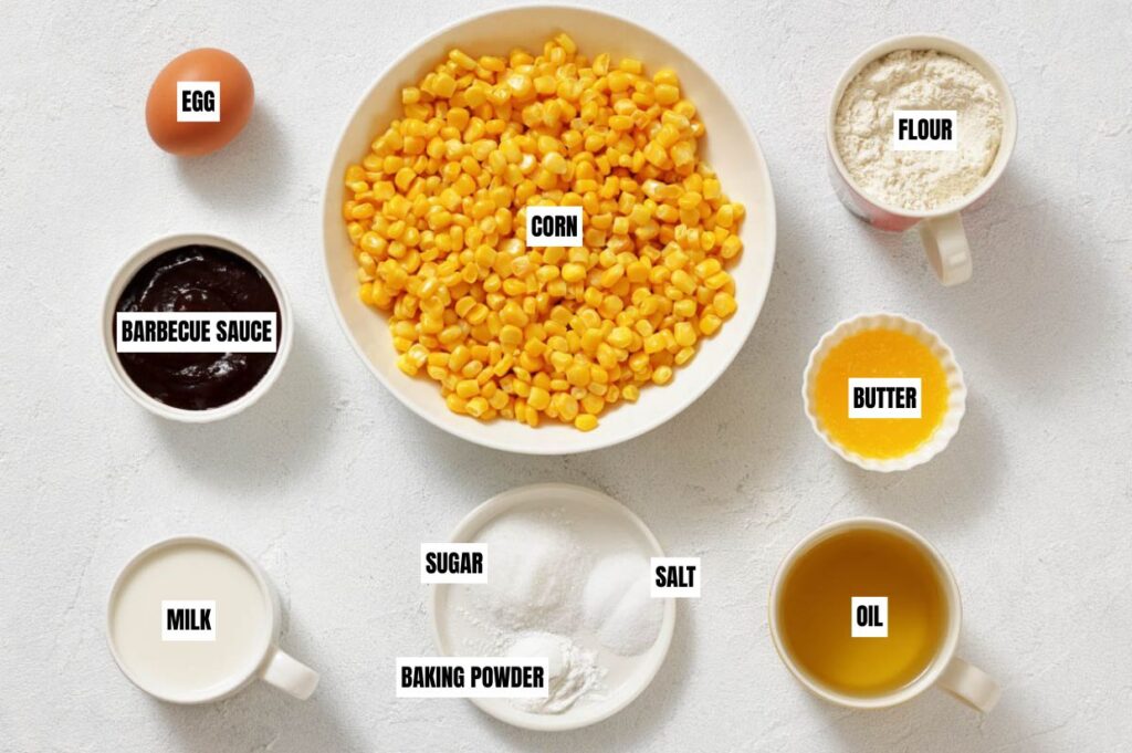 Overhead view of cooking ingredients on a white surface: corn, flour, egg, barbecue sauce, milk, sugar, salt, baking powder, butter, and oil.