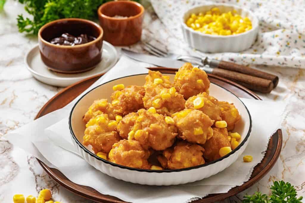 A plate of golden corn nuggets sprinkled with corn kernels, served on a wooden tray. Nearby, brown dipping sauce bowls and parsley add fresh accents.