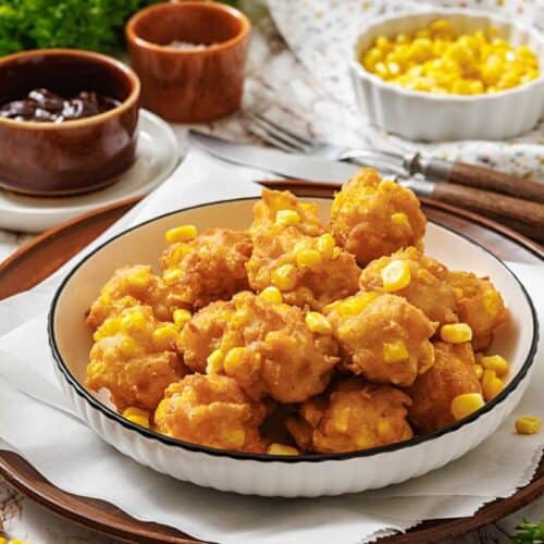 A plate of golden corn nuggets sprinkled with corn kernels, served on a wooden tray. Nearby, brown dipping sauce bowls and parsley add fresh accents.