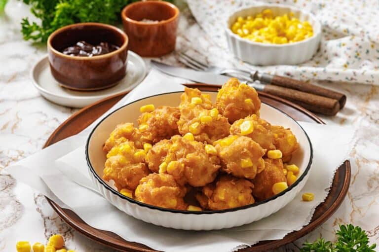 A plate of golden corn nuggets sprinkled with corn kernels, served on a wooden tray. Nearby, brown dipping sauce bowls and parsley add fresh accents.