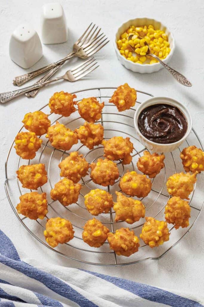 Golden corn fritters on a cooling rack with a small bowl of dark dipping sauce. Nearby are forks, salt, and a bowl of corn, creating a cozy setting.