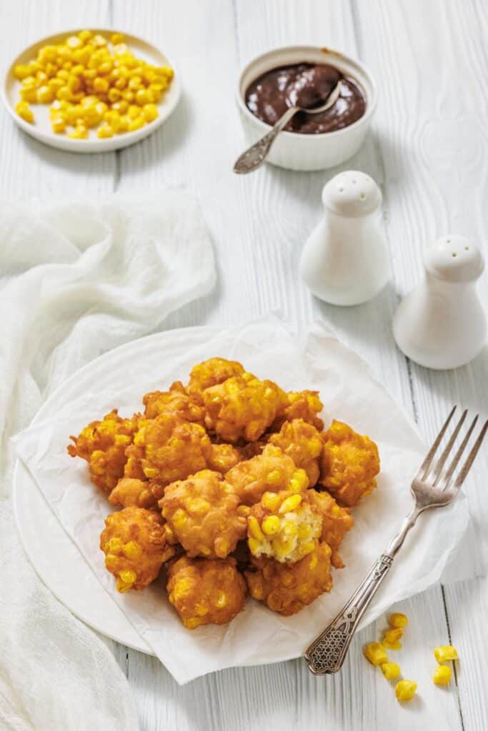 Golden corn fritters piled on a white plate, accompanied by a dish of chocolate sauce, corn kernels, and salt shakers. The setting feels cozy.