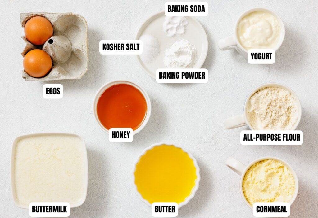 A collection of labeled baking ingredients on a white surface includes eggs, buttermilk, honey, butter, baking soda, baking powder, kosher salt, yogurt, all-purpose flour, and cornmeal.