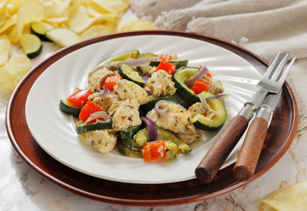 Plate of seasoned chicken, zucchini, red bell peppers, and red onions on a wooden-rimmed plate. Rustic backdrop with fork and knife. Cozy and appetizing.