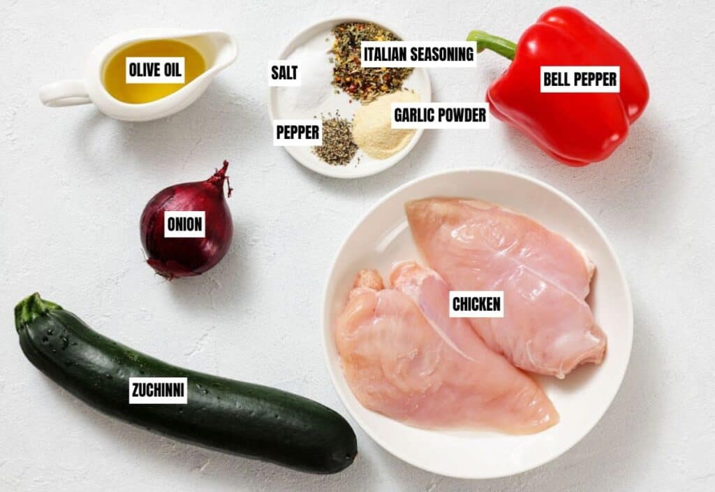 Raw ingredients for a dish on a white surface: chicken breasts on a plate, red bell pepper, red onion, zucchini, olive oil, and a plate with salt, pepper, Italian seasoning, and garlic powder.