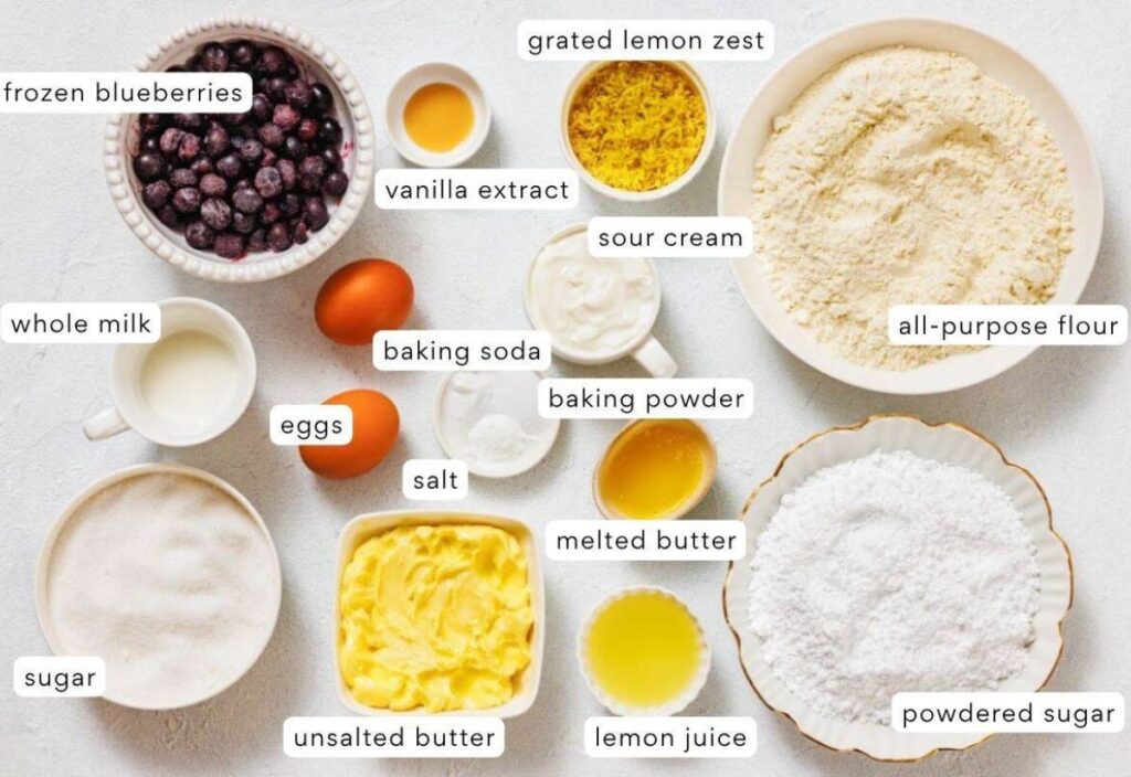A collection of baking essentials—blueberries, lemon zest, vanilla, sour cream, flour, milk, eggs, baking soda, baking powder, salt, butter, lemon juice, powdered sugar, and sugar—arranged in small bowls and containers, each clearly labeled for a blueberry lemon dessert recipe.