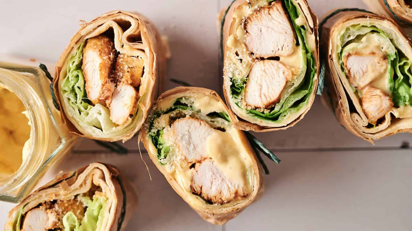 Close-up of sliced chicken wraps with lettuce, cheese, and a creamy sauce, next to an open jar of mustard on a wooden surface.
